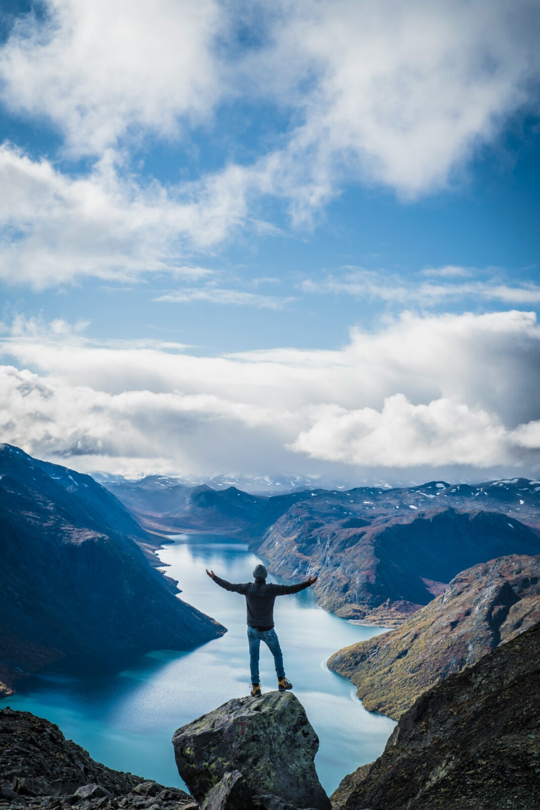 Hiker at the top of a precarious rock overlooking a beautiful lake and mountain valley on a clear day with hands held high in triumph.