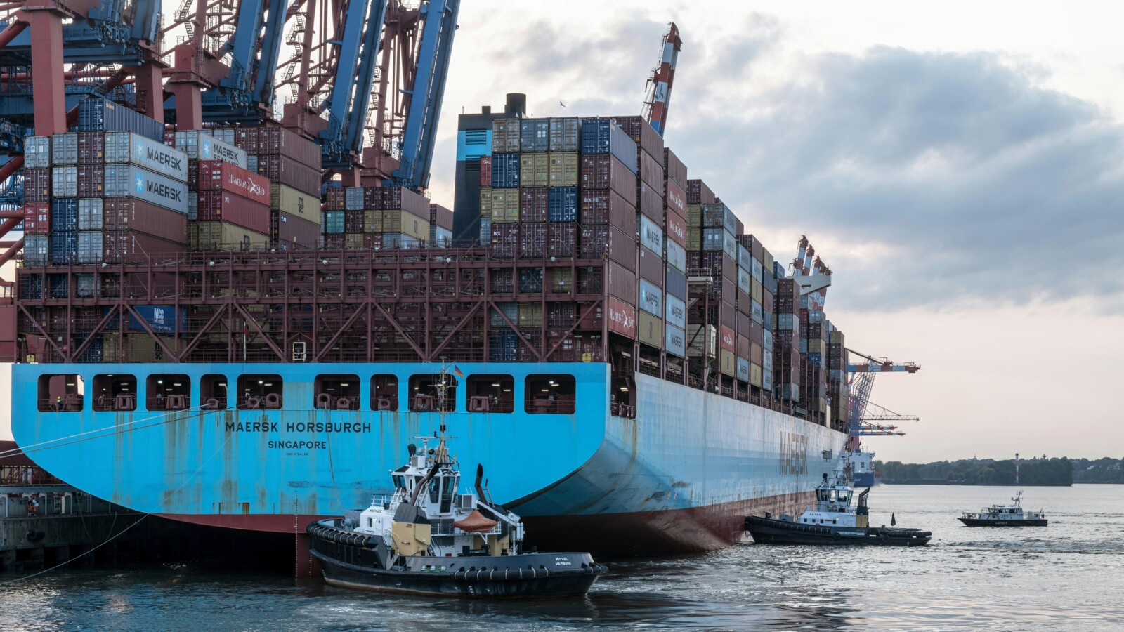 Large cargo ship Singapore docking at a port with hundreds of crates, pushed by two tugboats under a cloudy sky.